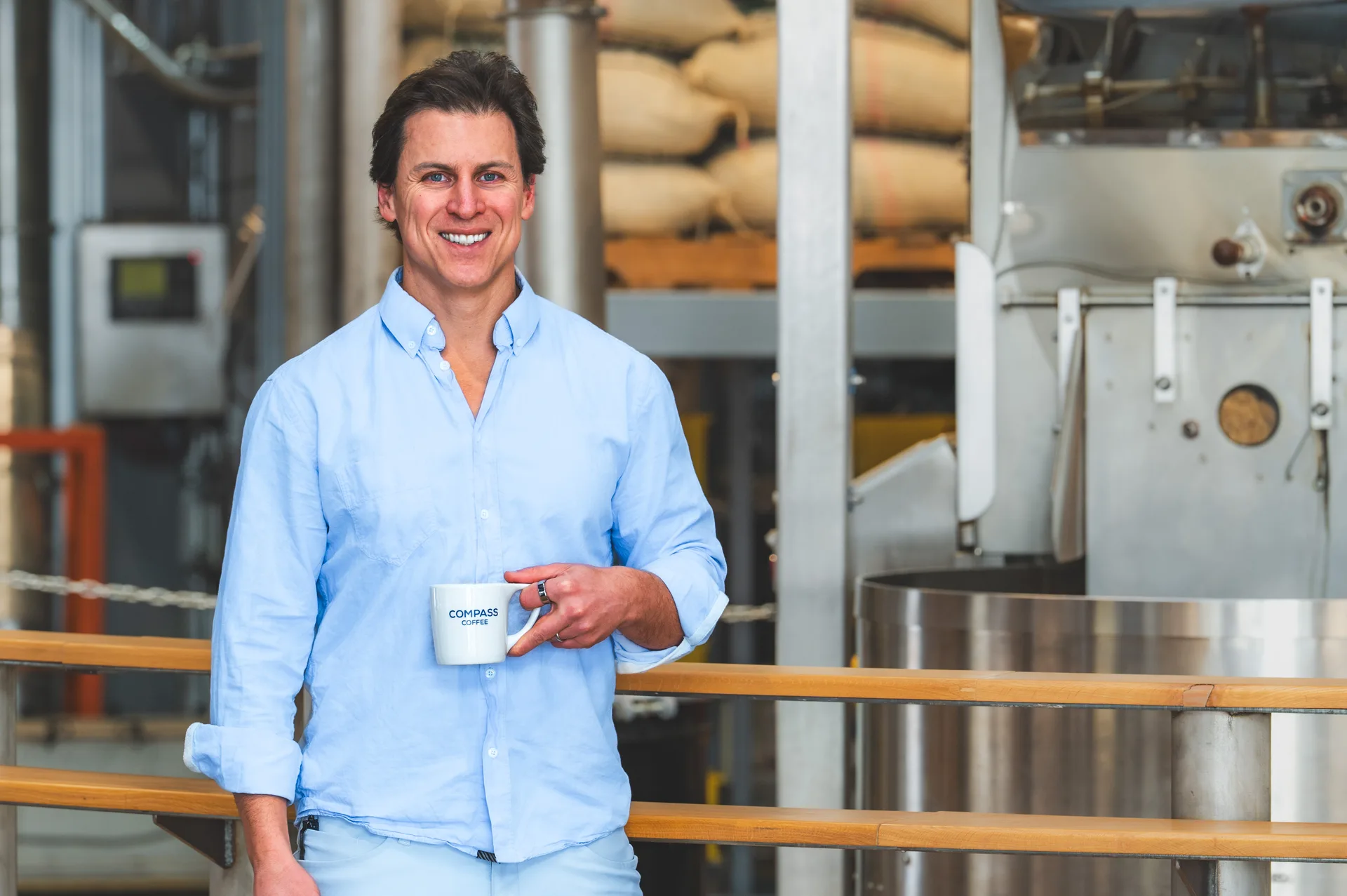 A man holding a Compass Coffee mug beside roasting equipment