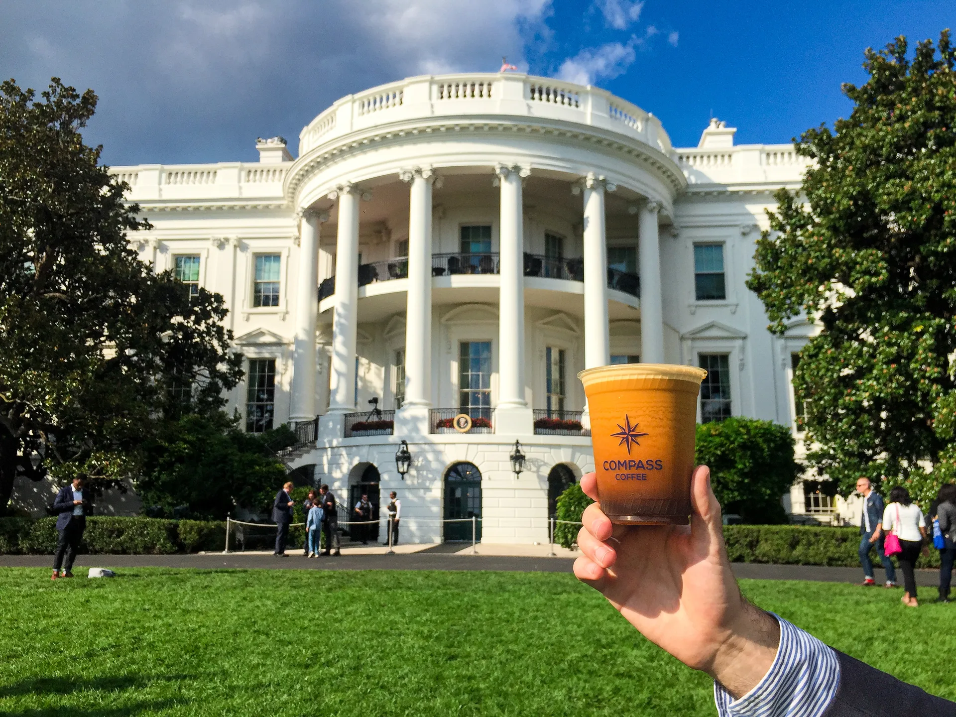A hand holding a Compass Coffee cup in front of the White House