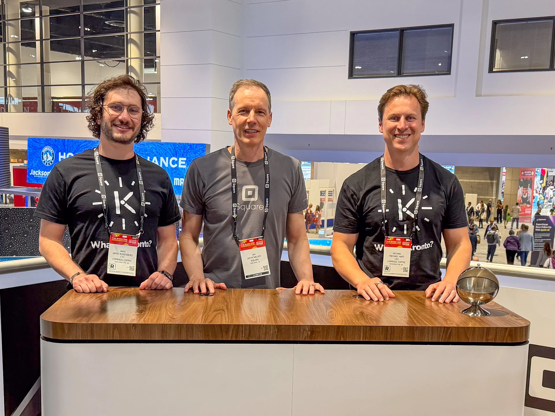 Three people wearing event badges stand behind a wooden counter in an exhibition hall