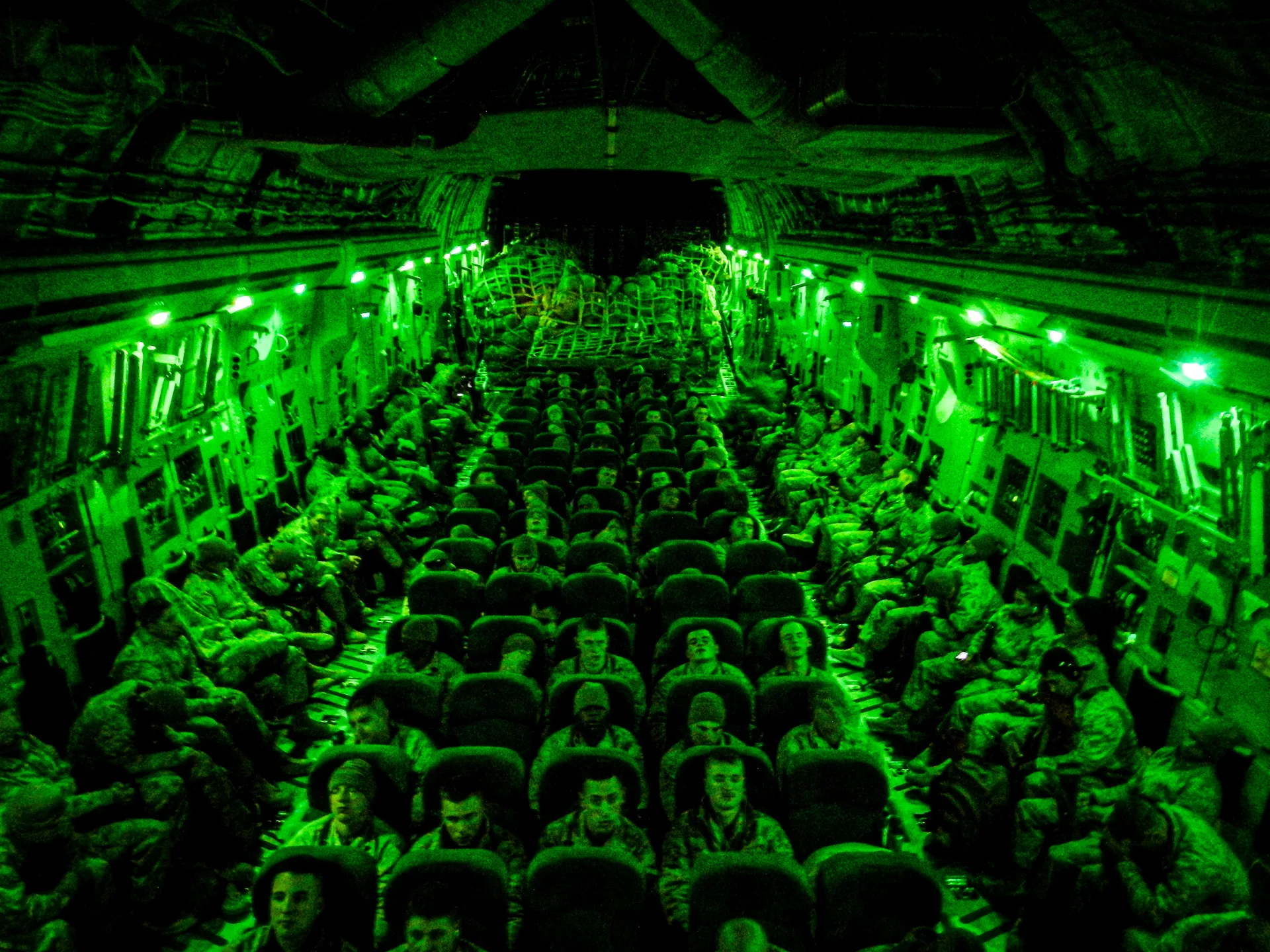 Service members seated inside a military transport aircraft under green cabin lighting